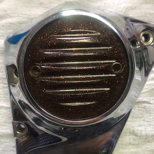 May include: Chrome motorcycle engine cover with a round, brown, glittery lens. The lens has a series of horizontal lines.