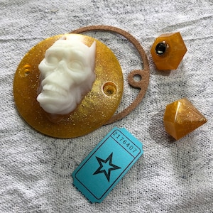 May include: A gold and white resin skull with a glitter finish. The skull is sitting on a gold glitter background. There are three gold glitter dice in the image. The dice are in the shape of a pyramid. The image also includes a blue ticket with the number 2176407 printed on it.