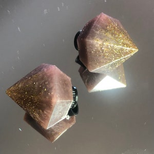 Peut inclure: Deux pinces à cheveux en forme de diamant rose avec des paillettes dorées incrustées dans la résine. Les pinces sont fixées à des barrettes en plastique noir.