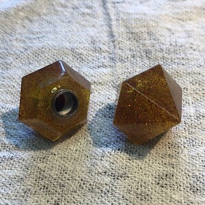 Può includere: Due pomelli geometrici in resina color oro, sfaccettati e con una finitura scintillante. Un pomello ha un foro filettato al centro.