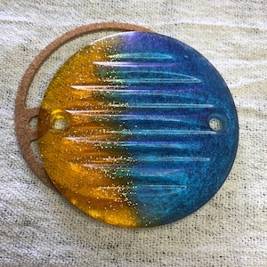 Puede incluir: Una tapa redonda de resina transparente con un acabado de purpurina dorada y azul iridiscente. La tapa tiene dos agujeros en el borde.