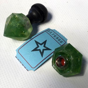 May include: Two green glass bottle stoppers with a star-shaped ticket with the number 2176988. One stopper has a black knob on top and the other has a clear opening.