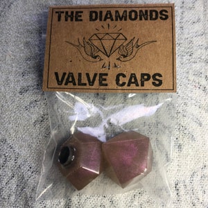 Puede incluir: Dos tapones de válvula en forma de diamante de color púrpura iridiscente, empaquetados en una bolsa de plástico transparente con una etiqueta de papel marrón que dice "THE DIAMONDS VALVE CAPS".