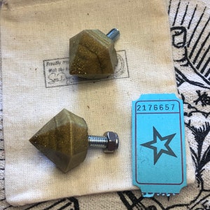 May include: Two gold-colored diamond-shaped drawer pulls with silver screws and a blue ticket with the number 2176657 and a star printed on it.