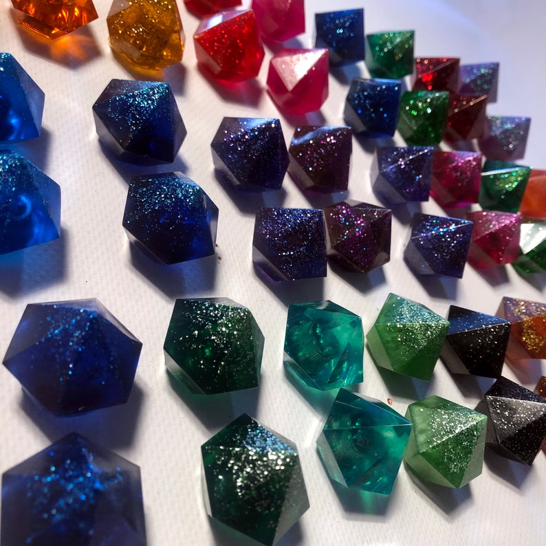 May include: A collection of colorful, glittery, resin dice in various shades of blue, green, red, and purple. The dice are shaped like a pentagon and have a sparkly finish.