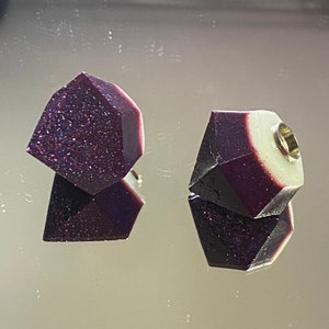 May include: Two purple and white resin gemstone earrings with a gold accent. The earrings are shaped like a faceted gem.