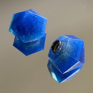 Peut inclure: Deux bouchons d'oreille en résine géométrique bleue et pailletée. Les bouchons sont en forme de gemme multi-facettes.