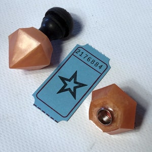 May include: Two peach-colored, faceted, geometric bottle stoppers with a threaded metal insert. One stopper is attached to a black wooden knob. A blue ticket with a black star and the number 2176884 is in the background.