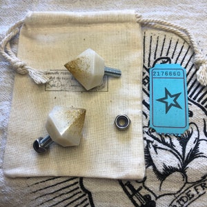 May include: Two white and gold glitter geometric knobs with bolts and nuts, next to a small bearing and a drawstring bag. A blue ticket with a star and the number 2176660 is also in the image.