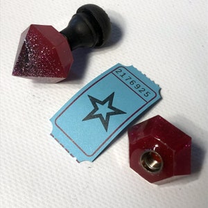 Puede incluir: Dos tapones de botella facetados rojos y brillantes. Uno tiene forma de diamante y el otro es un hexágono. El tapón hexagonal tiene un agujero roscado en el centro. Un billete azul con una estrella y el número 2176925 está en el fondo.