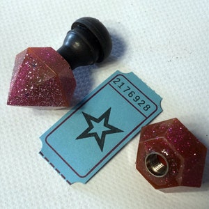 Puede incluir: Dos tapones de botella de purpurina rosa con un pomo negro en la parte superior. Un tapón tiene forma de diamante y el otro tiene forma de hexágono. Un billete azul con una estrella y el número 2176928 está en el fondo.