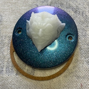 以下が含まれることがあります： 丸いターコイズブルーの金属製のプレートの上に、白い3D彫刻の悪魔の頭が乗っています。プレートには側面に2つの穴があり、茶色の革のリングで囲まれています。