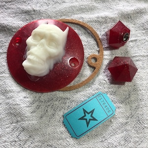 May include: A red round resin coaster with a white skull sculpture on top. The coaster has a glittery finish. There are two red resin geometric shapes and a blue ticket with a star on it.