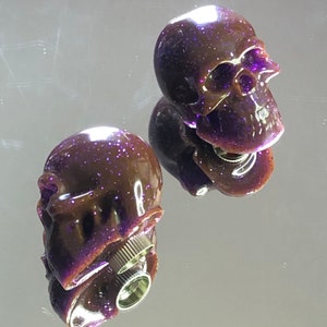 Könnte beinhalten: Zwei violette und braune, schädelförmige Schaltknöpfe mit glitzernder Oberfläche. Die Knöpfe haben einen Gewinde-Metallfuß.