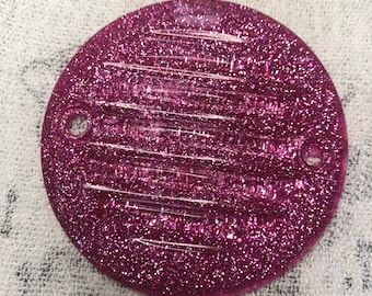 Puede incluir: Un disco redondo de resina rosa cubierto de purpurina con dos pequeños agujeros. El disco tiene un patrón texturizado a rayas.