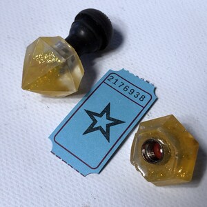 May include: Two clear glass bottle stoppers with gold glitter. One stopper has a black wooden knob on top. The other stopper has a red metal cap. A blue ticket with a black star and the number 2176938 is in the background.