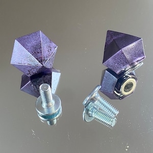 Peut inclure: Deux poignées de tiroir en forme de diamant violet avec des vis argentées. Les poignées de tiroir ont une surface texturée et sont fixées à des plaques en métal argenté.