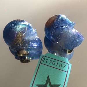 Könnte beinhalten: Zwei blaue und schillernde Totenkopf-Flaschenverschlüsse aus Harz mit einem goldenen Metallring. Die Verschlüsse befinden sich auf einer blauen Oberfläche mit einem türkisfarbenen Ticket, auf dem die Nummer 2176107 aufgedruckt ist.