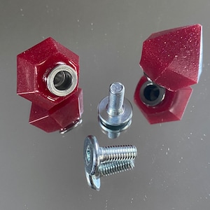 Peut inclure: Gros plan de plusieurs boutons rouges, facettés, de forme géométrique, avec des inserts en métal argenté et des vis argentées assorties. Les boutons ont un aspect brillant et translucide. Les vis sont de tailles variables.