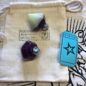 Pode incluir: Dois pequenos objetos em forma de diamante de resina brilhante com bases de metal rosqueadas. Os diamantes são roxos e verdes com uma base branca. Os objetos estão em uma bolsa de tecido branco com um carimbo que diz "Apresentado com orgulho... com o coração e a alma de..."