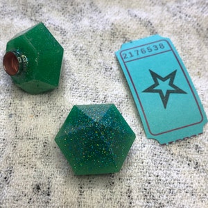 Può includere: Due dadi geometrici in resina verde e scintillante con un biglietto a forma di stella su cui è stampato il numero 2176538.