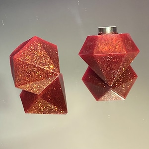 May include: Two red, glittery, geometric earrings with a faceted design. The earrings are made of resin and have a gold glitter finish.