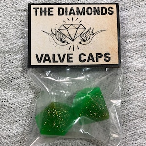 Może przedstawiać: Dwa zielone korki do zaworów z złotym brokatem. Korki są zapakowane w przezroczysty plastikowy woreczek z etykietą z napisem "The Diamonds Valve Caps".