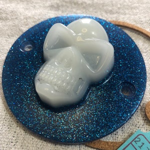 Peut inclure: Un crâne blanc sur un fond de paillettes bleues. Le crâne est en résine et a une finition brillante. Le fond est bleu foncé avec une texture de paillettes fines.