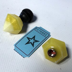 May include: Two yellow, faceted, geometric bottle stoppers with a black knob on one. The stoppers are on a white surface with a blue ticket with a star and the number 2176881.
