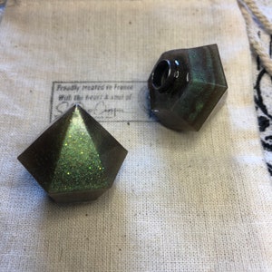 May include: Two green and black resin bottle stoppers with glitter. One stopper is a pyramid shape and the other is a rounded shape with a hole in the top.