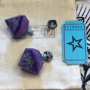 Peut inclure: Deux tirettes de tiroir en résine de forme diamantée, de couleur violette avec des paillettes argentées et une vis argentée. Les tirettes sont sur un fond de tissu blanc.