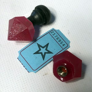 Puede incluir: Dos tapones de botella de resina en forma de diamante rojo y brillante con un pomo negro en uno. Un billete azul con una estrella y el número 2176954 está en el fondo.