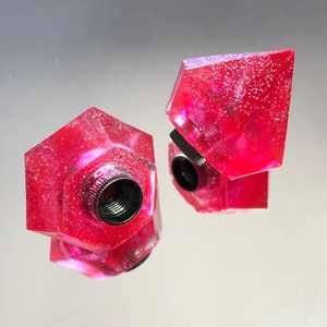 Puede incluir: Dos tapones de botella de resina rosa y brillante con un inserto roscado de metal. Un tapón tiene forma de diamante y el otro tiene forma de hexágono.