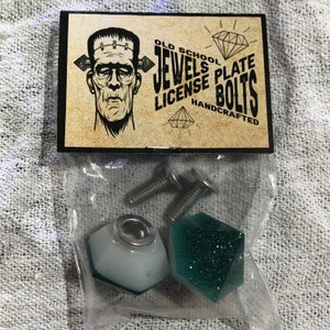 May include: A set of two green and white diamond-shaped skateboard license plate bolts with screws. The packaging includes a sticker with the text "Old School Jewels Plate License Bolts Handcrafted".