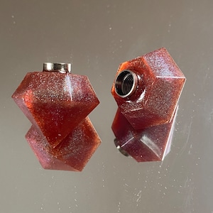 Peut inclure: Deux bouchons de bouteille en résine rouge et pailletée, de forme géométrique, avec un insert en métal argenté.