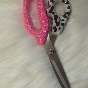 Puede incluir: Tijeras con un mango rosa brillante y un mango con un diseño de manchas blancas y negras. Las cuchillas son plateadas. Las tijeras están sobre una superficie blanca y esponjosa.
