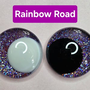 Puede incluir: Dos cabujones redondos de vidrio brillante con el texto "Rainbow Road" sobre un fondo morado. Un cabujón tiene un ojo blanco con un reflejo en forma de media luna, y el otro tiene un ojo negro con un reflejo en forma de media luna.