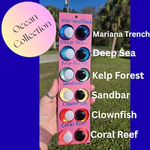 May include: A pink card displaying six pairs of craft eyes with an ocean theme. Each pair has a white pupil and a coloured iris, labelled "Mariana Trench," "Deep Sea," "Kelp Forest," "Sandbar," "Clownfish," and "Coral Reef." The words "Ocean Collection" are in a circle.