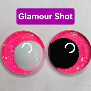 May include: Two round, pink-rimmed eye cabochons with glitter. One has a white iris, the other a black iris. Both have a white crescent-shaped highlight. The text "Glamour Shot" is in a purple rectangle above.