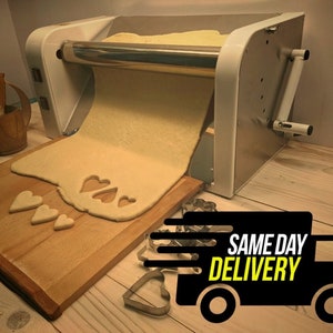 May include: A white electric dough sheeter with a stainless steel roller, rolling out a sheet of dough on a wooden cutting board.  Heart-shaped cookie cutters are scattered on the board.  The text "SAME DAY DELIVERY" is on a black truck icon.