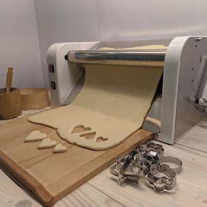 May include: A white electric dough roller with a stainless steel roller and a crank handle. A sheet of dough is being rolled out of the machine onto a wooden cutting board. There are heart-shaped cookie cutters and a few heart-shaped cookies on the cutting board.