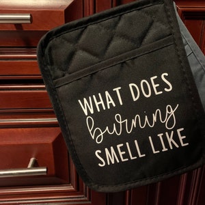 May include: Black quilted oven mitt with the text "WHAT DOES burning SMELL LIKE" in white script. The mitt has a pocket and a loop for hanging. The background shows a dark wood cabinet.