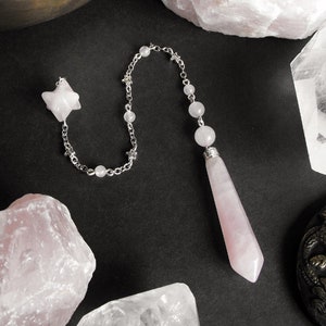 Può includere: Un pendolo di quarzo rosa rosa con una catena e perline argentate, circondato da cristalli di quarzo rosa e quarzo trasparente su uno sfondo nero.