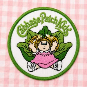 May include: A circular embroidered patch featuring a Cabbage Patch Kids logo in green text above a cartoon character with blonde hair in pigtails, wearing a pink dress and white shoes, positioned in front of large green cabbage leaves.