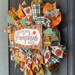 May include: A decorative wreath featuring orange, green, and tan ribbons with pumpkin and leaf patterns. A rectangular wooden sign in the center displays the text "AUTUMN LEAVES AND PUMPKINS PLEASE" with a small orange pumpkin icon on a diamond background.