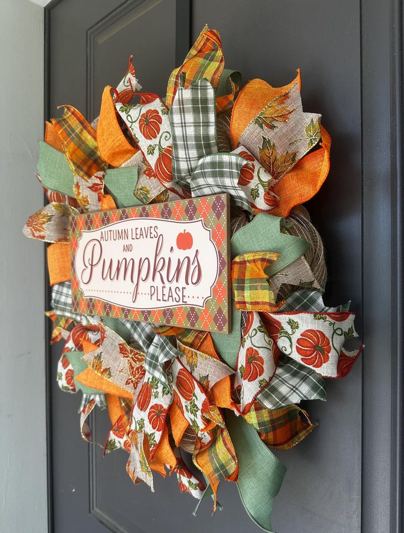 May include: A decorative wreath featuring orange, green, and tan ribbons with pumpkin and leaf patterns. A rectangular wooden sign in the center displays the text "AUTUMN LEAVES AND PUMPKINS PLEASE" with a small orange pumpkin icon on a diamond background.