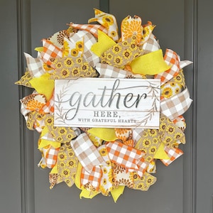 May include: A decorative wreath featuring a central rectangular sign with the text gather HERE, WITH GRATEFUL HEARTS. The wreath is constructed from layers of yellow, orange, and white plaid ribbons, along with ribbons patterned with sunflowers.