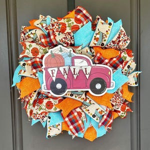 May include: A decorative wreath featuring a central cutout of a pink truck carrying pumpkins with a banner that reads FALL. The wreath is constructed from layers of mesh and fabric ribbons in orange, turquoise, and plaid patterns with pumpkin prints.