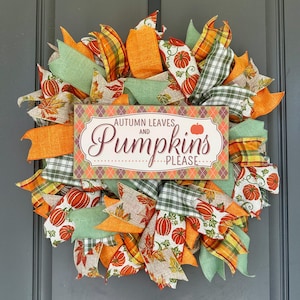 May include: A decorative wreath featuring orange, green, and plaid ribbons with pumpkin and leaf patterns. A rectangular sign in the center displays the text "AUTUMN LEAVES AND Pumpkins PLEASE" against a diamond-patterned background.