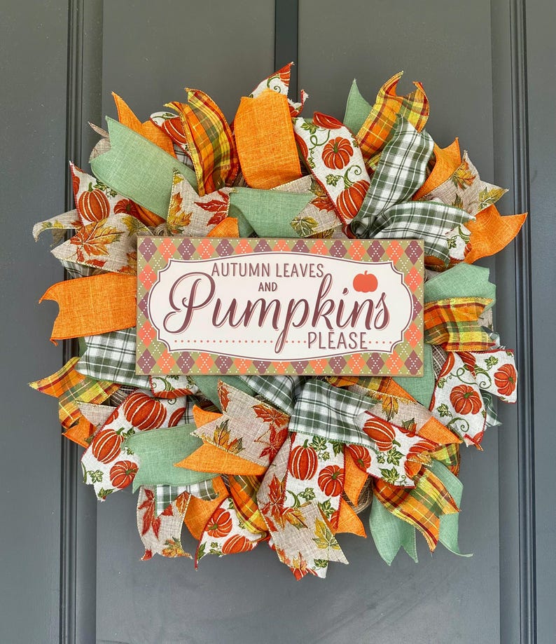 May include: A decorative wreath featuring orange, green, and plaid ribbons with pumpkin and leaf patterns. A rectangular sign in the center displays the text "AUTUMN LEAVES AND Pumpkins PLEASE" against a diamond-patterned background.